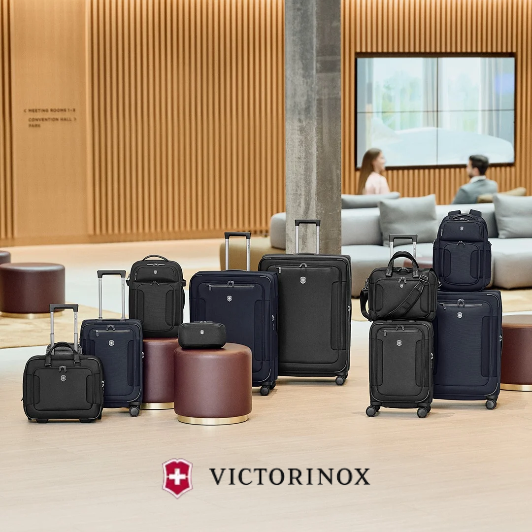A collection of Victorinox rolling luggage and backpacks in black, navy, and tan, arranged on a polished wood floor in a modern hotel lobby. The Victorinox logo is displayed at the bottom of the image.